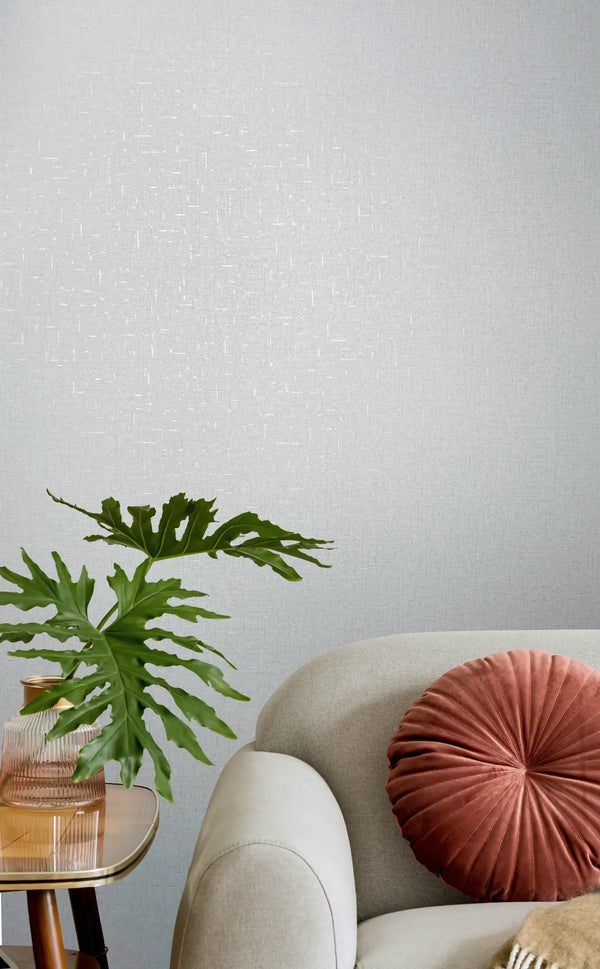 Marburg City Romance 35116 Light gray textured wallpaper behind a light upholstered sofa with a round rust-colored cushion, a small side table, and a leafy green plant in a ribbed glass vase.