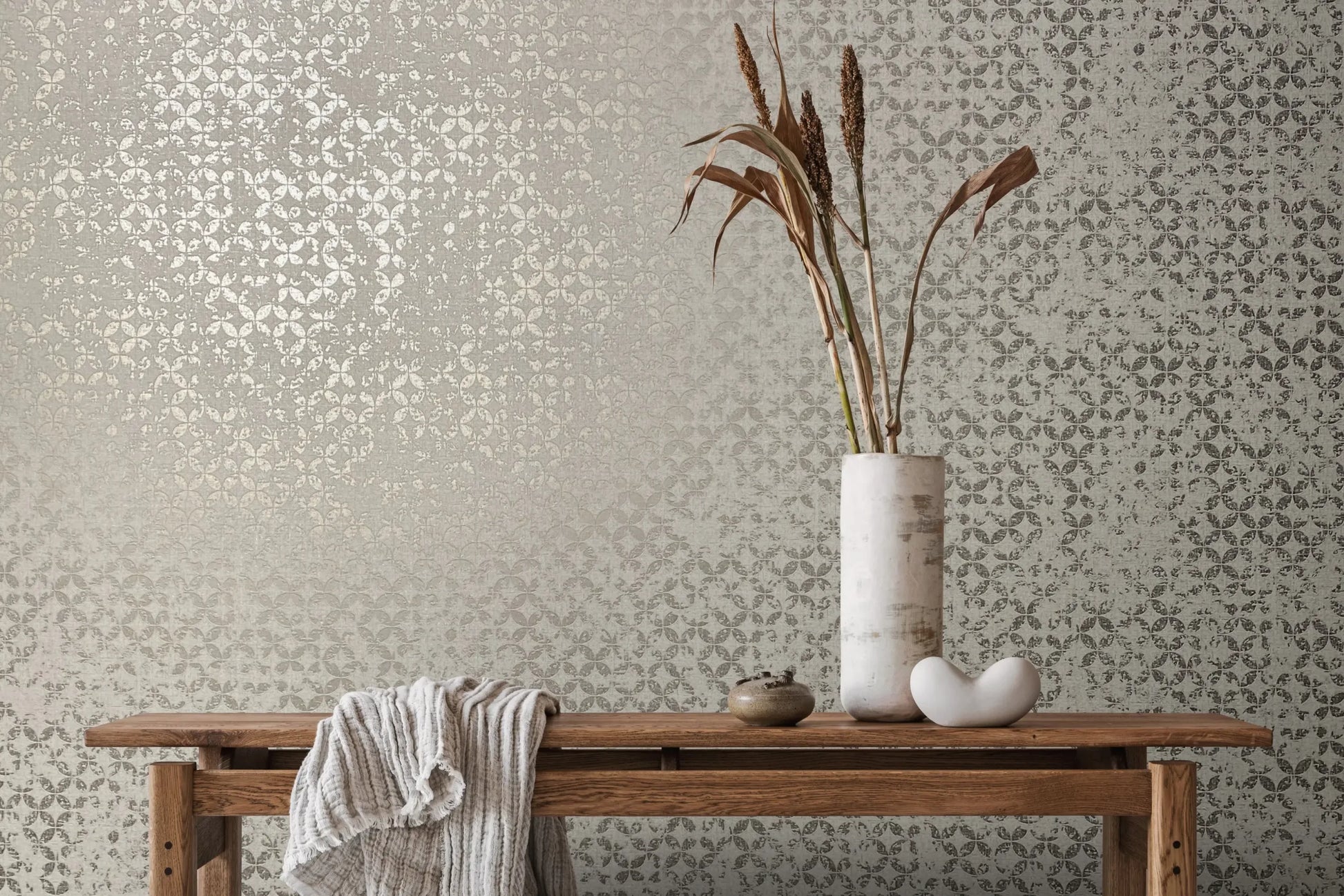 Marburg City Romance 35118 Gray wallpaper with a small repeating petal/clover pattern and a subtle sheen, shown behind a wooden bench styled with a draped textured throw, a tall white cylindrical vase holding dried stems, a small round jar, and a white sculptural object.