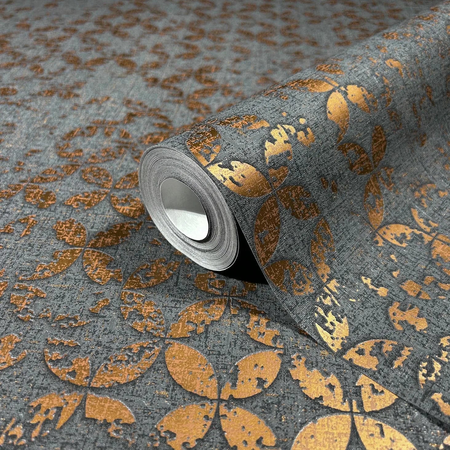 Marburg City Romance 35119 Close-up of a wallpaper roll on a matching surface showing a dark gray textured background with metallic gold petal-like geometric shapes and a distressed finish.