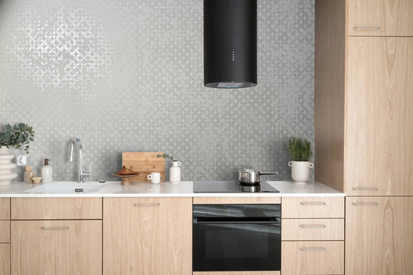 Marburg City Romance 35121 Kitchen scene with light wood cabinets and a light gray leaf-pattern wallpaper with a subtle sheen. A black cylindrical range hood hangs above a cooktop with a saucepan, with an oven below. On the countertop are a sink and faucet, cutting board, mugs, a soap/l.