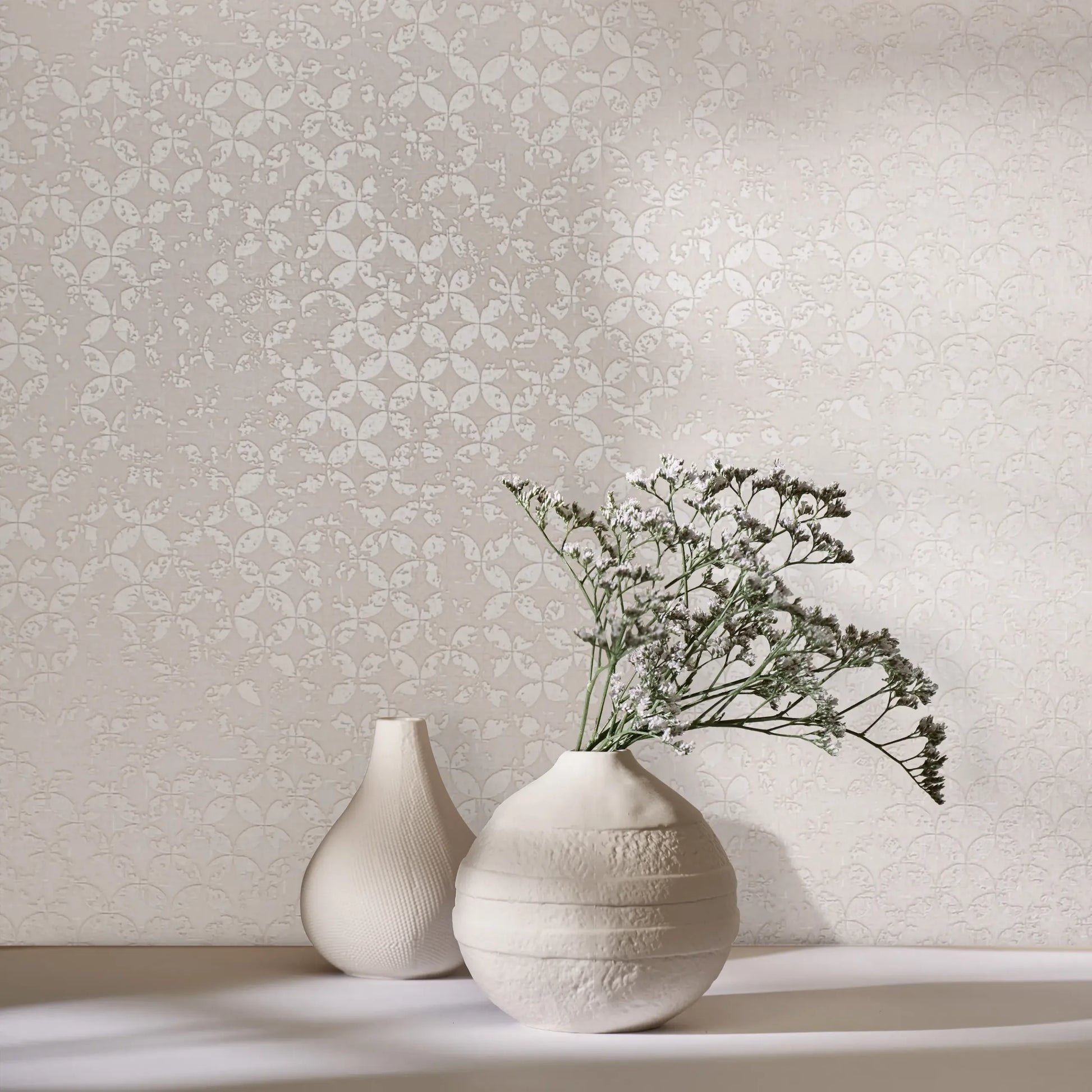 Marburg City Romance 35122 Light beige wallpaper featuring a small repeating rosette-style pattern. In front, two ceramic vases sit on a surface, one holding delicate white flowers with soft shadows.