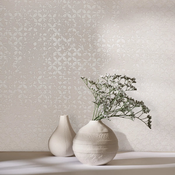 Marburg City Romance 35122 Light beige wallpaper featuring a small repeating rosette-style pattern. In front, two ceramic vases sit on a surface, one holding delicate white flowers with soft shadows.