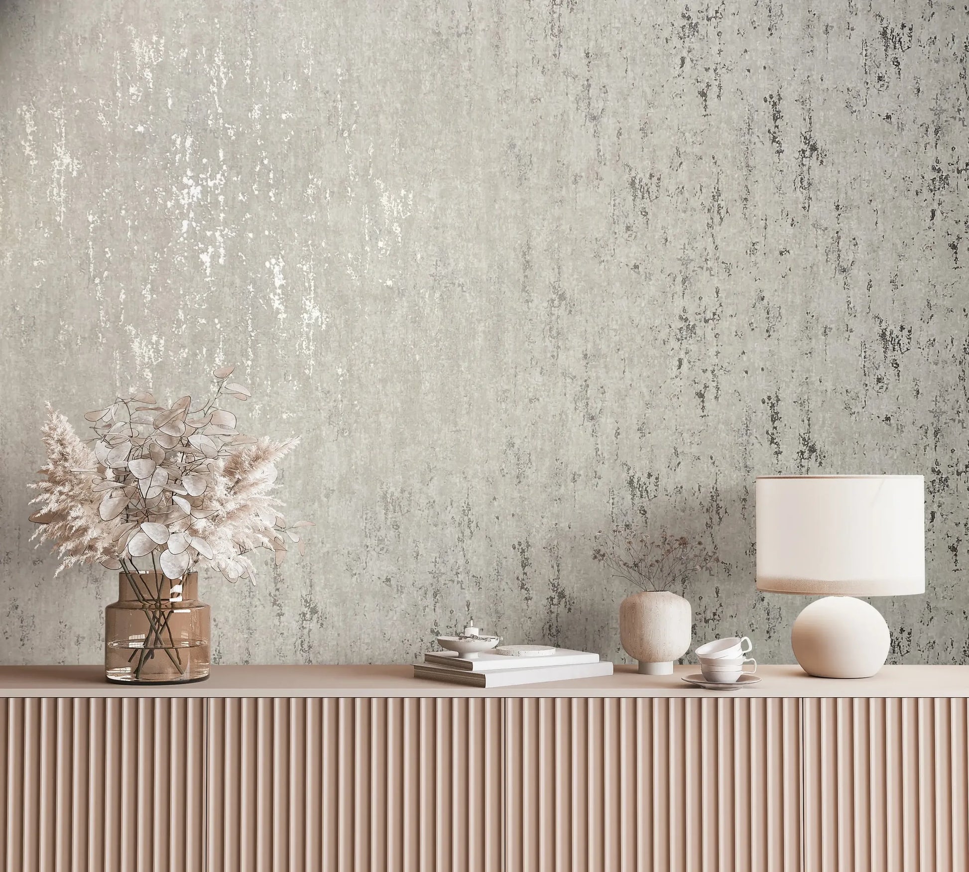 Marburg City Romance 35123 Styled scene showing gray distressed-texture wallpaper behind a fluted sideboard with a table lamp, a glass vase of dried florals, a small ceramic vase, stacked books, a teacup with saucer, and a small dish.