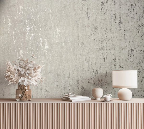 Marburg City Romance 35123 Styled scene showing gray distressed-texture wallpaper behind a fluted sideboard with a table lamp, a glass vase of dried florals, a small ceramic vase, stacked books, a teacup with saucer, and a small dish.
