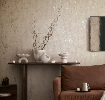 Marburg City Romance 35124 Beige mottled wallpaper with subtle metallic flecks behind a dark wood console styled with white ceramic vessels and a branch arrangement, adjacent to a brown sofa and a framed abstract artwork.