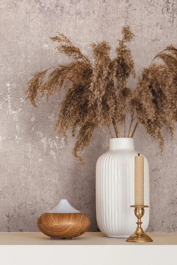 Marburg City Romance 35125 Close-up of textured, mottled plaster-effect wallpaper behind a ribbed white vase holding dried grasses, a brass candlestick with a taper, and a small wood-look diffuser on a shelf.