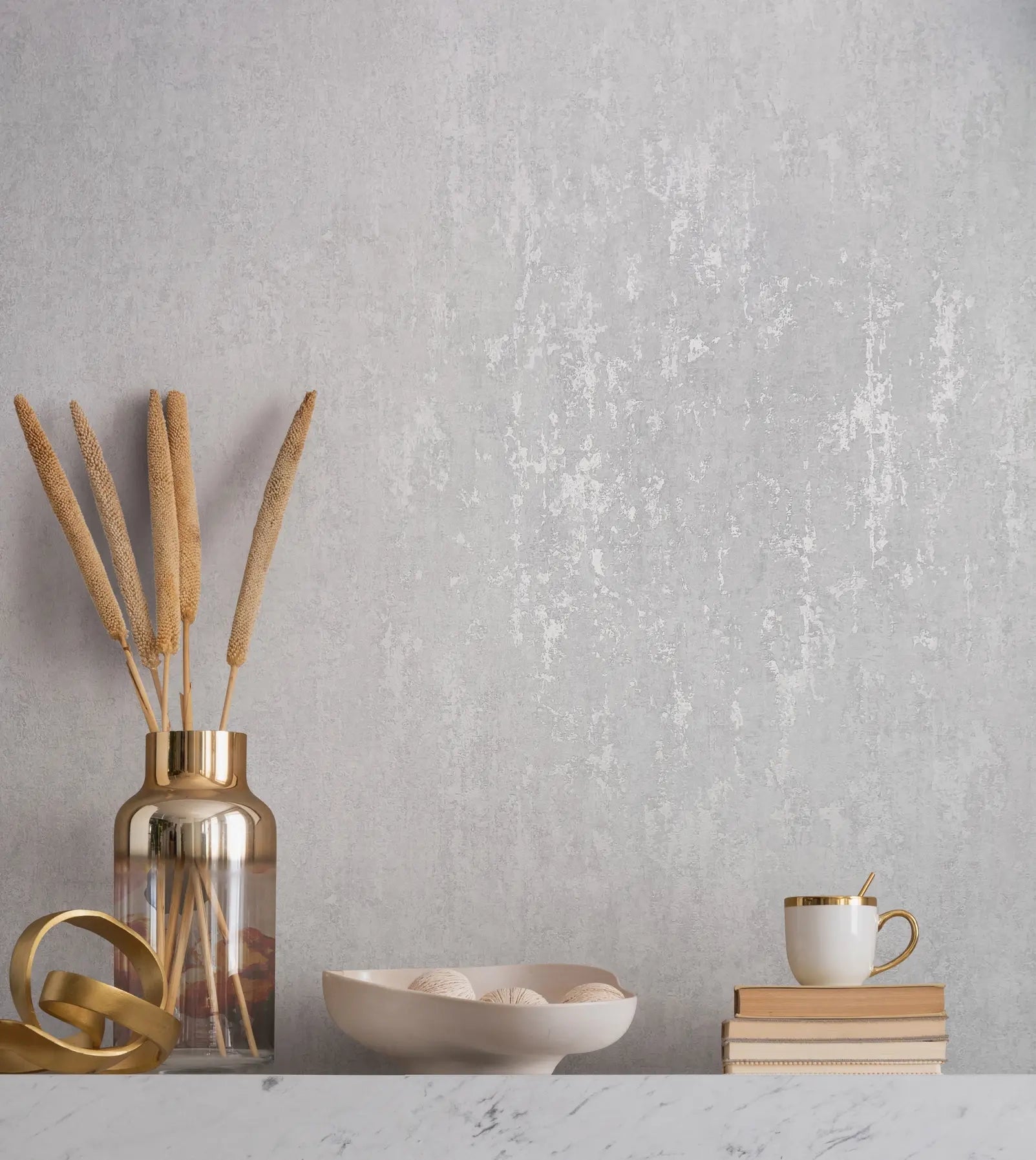 Marburg City Romance 35126 Light gray textured wallpaper with a weathered, mottled finish behind a marble surface styled with a glass vase holding dried stems, a metal ribbon decor piece, a shallow bowl with decorative balls, stacked books, and a white mug with a gold rim and spoon.