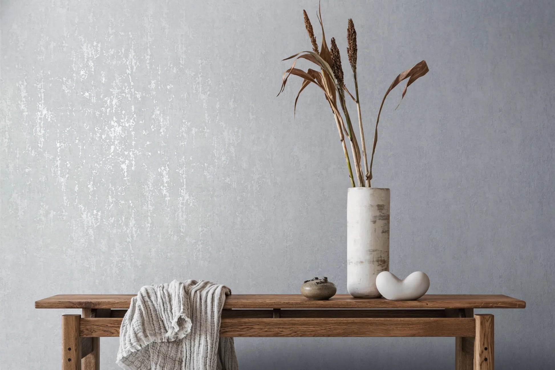 Marburg City Romance 35126 Light grey textured wallpaper behind a wooden bench styled with a tall cylindrical vase holding dried stems, a small ceramic jar, a white sculptural object, and a draped throw.