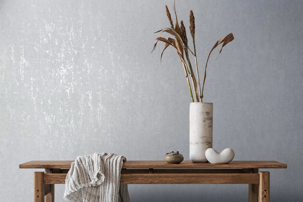 Marburg City Romance 35126 Light grey textured wallpaper behind a wooden bench styled with a tall cylindrical vase holding dried stems, a small ceramic jar, a white sculptural object, and a draped throw.