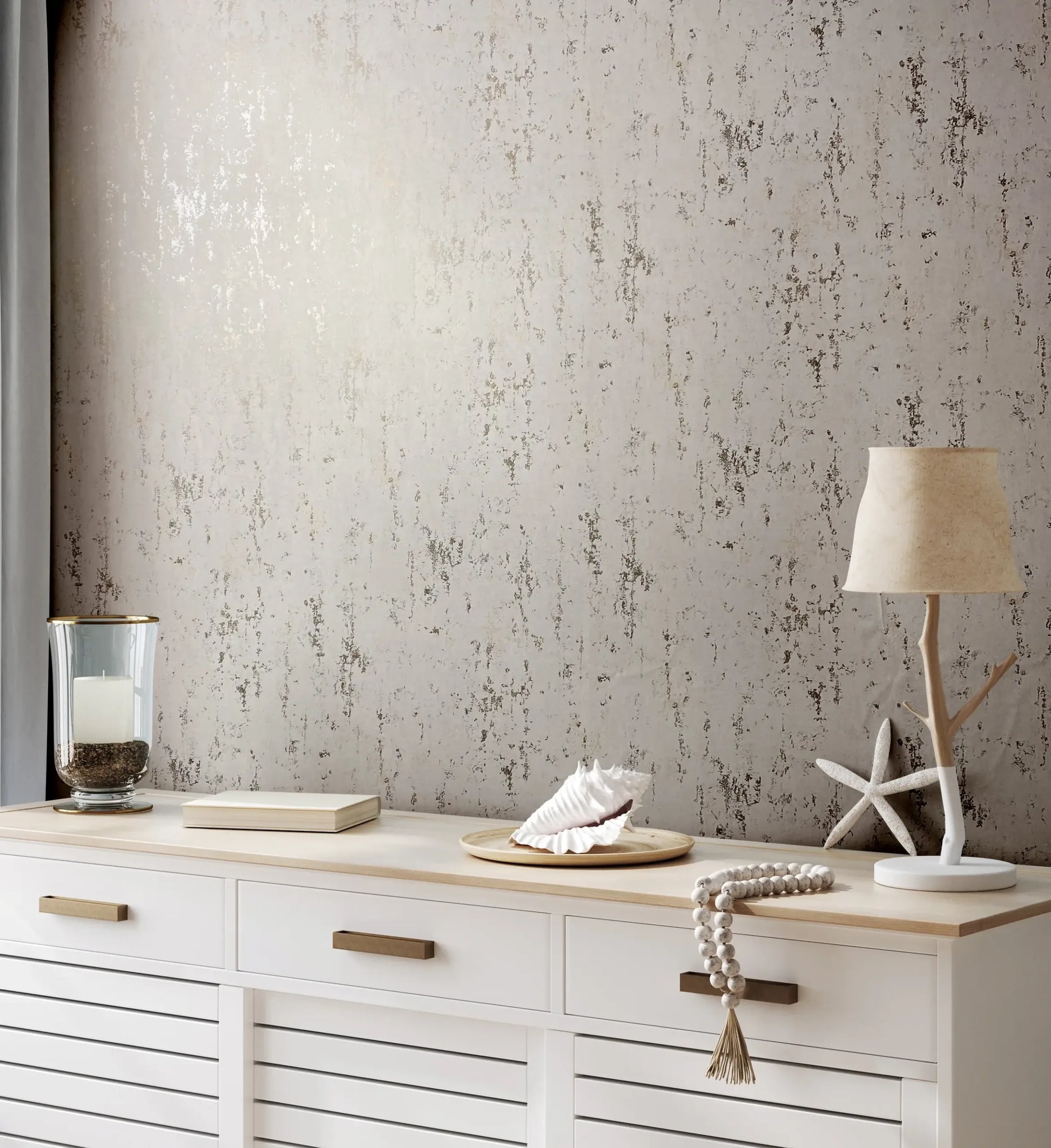 Marburg City Romance 35127 Light beige distressed-texture wallpaper behind a white sideboard styled with a table lamp, starfish decor, a seashell on a tray, a candle in a glass holder, and a book.