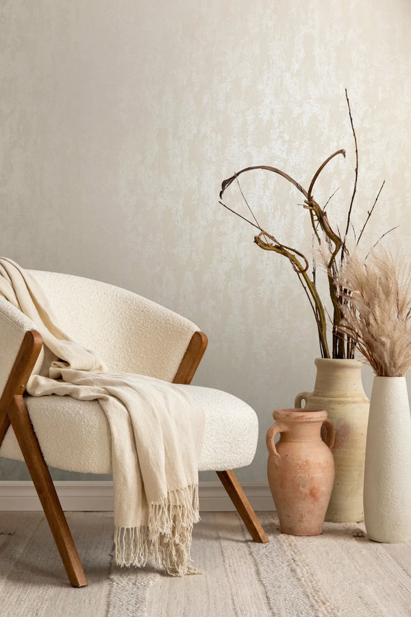 Marburg City Romance 35128 Beige textured effect wallpaper behind a cream upholstered chair with a draped throw, next to ceramic vases holding dried branches and dried grasses.