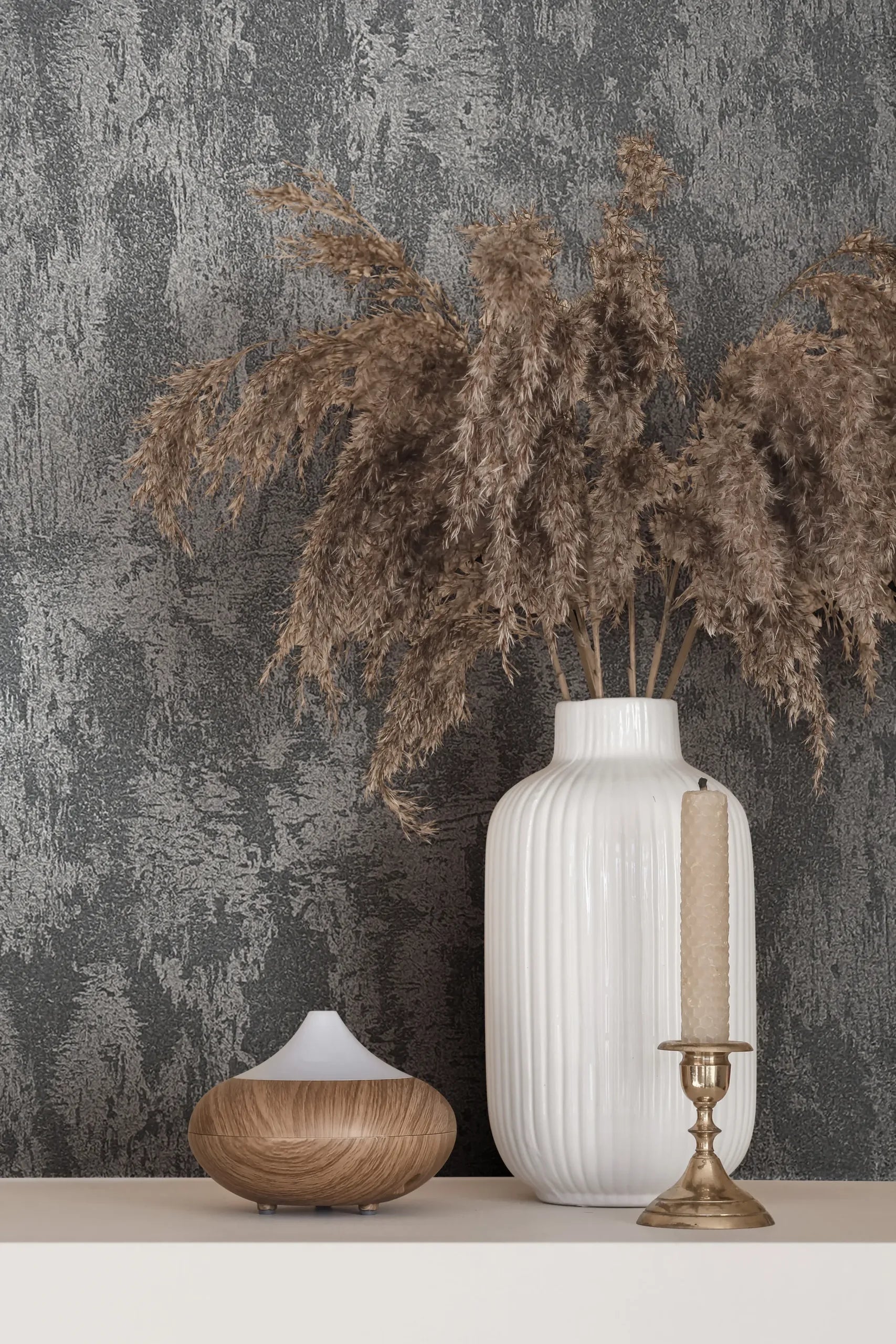 Marburg City Romance 35130 Textured grey wallpaper with an abstract plaster-like pattern behind a white ribbed vase holding dried grasses, a brass candlestick with a tapered candle, and a small wood-grain diffuser on a light shelf.
