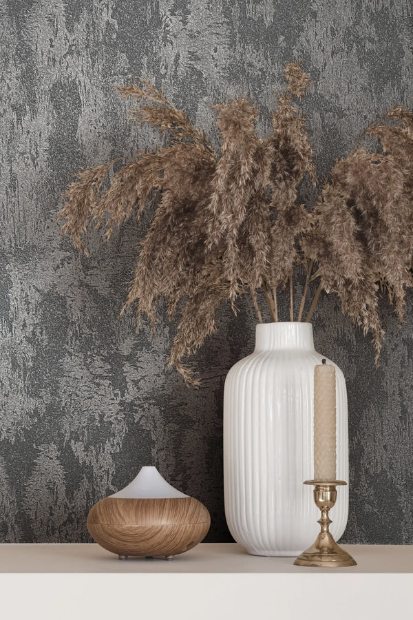 Marburg City Romance 35130 Textured grey wallpaper with an abstract plaster-like pattern behind a white ribbed vase holding dried grasses, a brass candlestick with a tapered candle, and a small wood-grain diffuser on a light shelf.