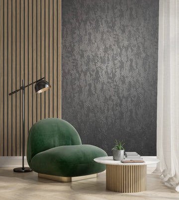 Marburg City Romance 35130 Dark gray abstract textured wallpaper on a feature wall behind a green lounge chair, with a round coffee table holding a small potted plant and books, a floor lamp, wood slat paneling, and a sheer curtain.