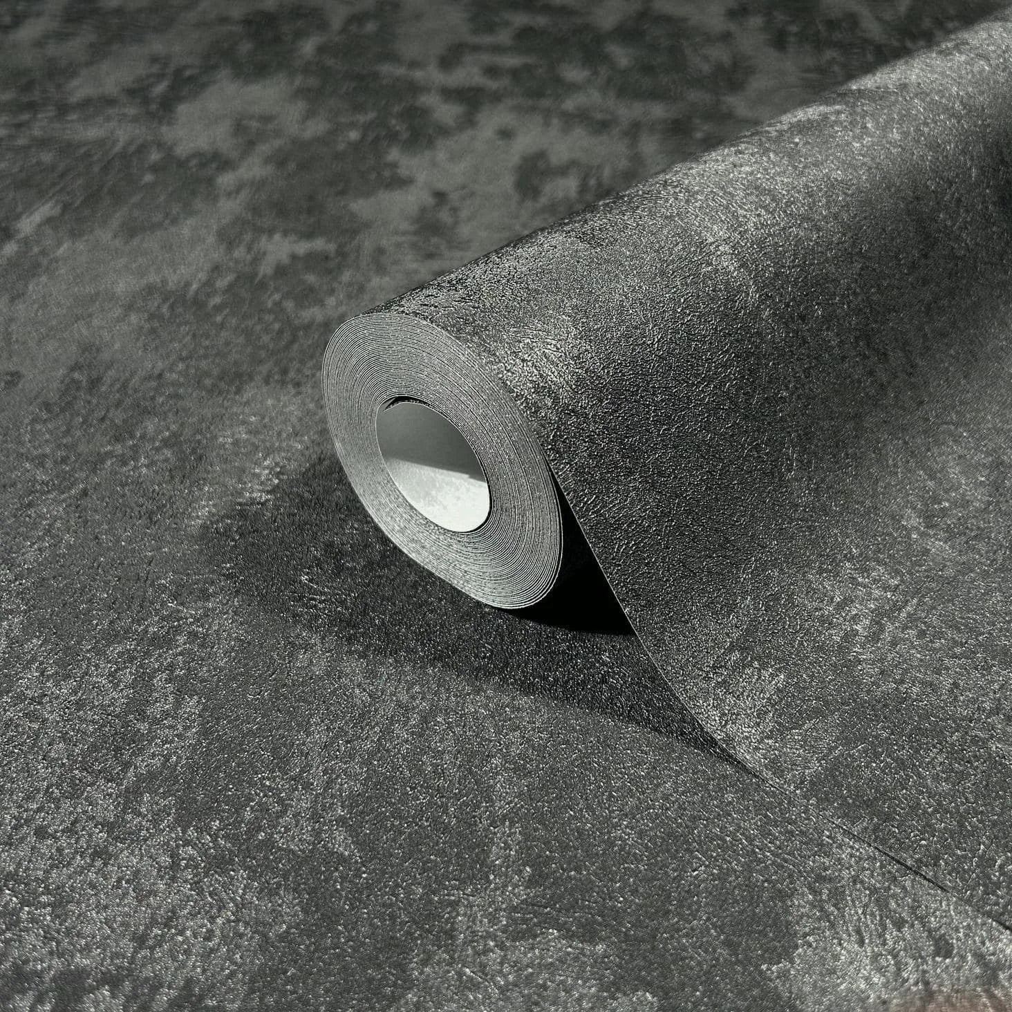 Marburg City Romance 35130 Close-up of a partially unrolled wallpaper roll resting on the same dark gray mottled textured wallpaper, showing a subtle sheen.
