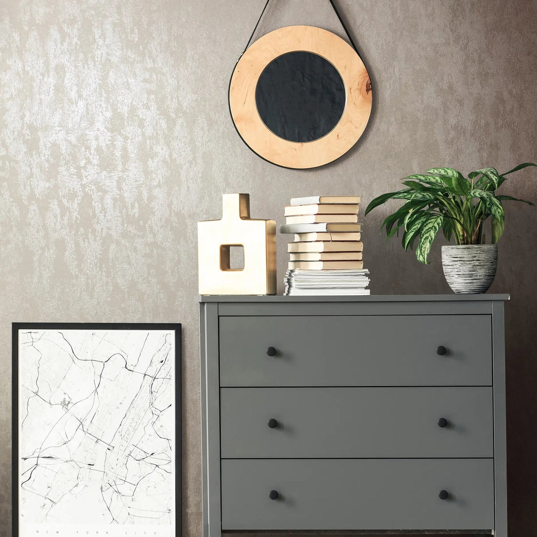 Marburg 35133 wallpaper lounge feature wall behind a grey three-drawer dresser with a round hanging mirror, stacked books, a sculptural vase, a potted plant, and a leaning city map print, modern calm mood.