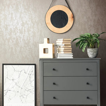 Marburg 35133 wallpaper lounge feature wall behind a grey three-drawer dresser with a round hanging mirror, stacked books, a sculptural vase, a potted plant, and a leaning city map print, modern calm mood.