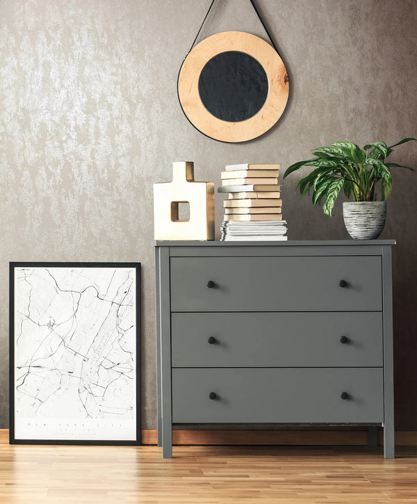 Marburg 35133 wallpaper lounge feature wall behind a grey three-drawer dresser with a round hanging mirror, stacked books, a sculptural vase, a potted plant, and a leaning city map print, modern calm mood.