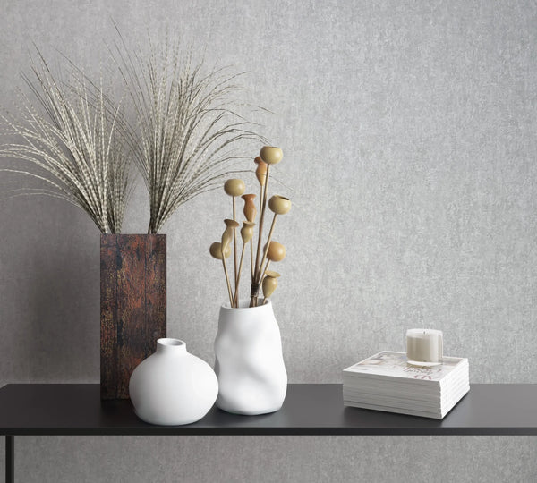 Marburg 35137 wallpaper lounge feature wall with light grey textured finish behind a black console table styled with white vases, a tall brown vase with grasses, stacked books and a candle, calm minimalist.