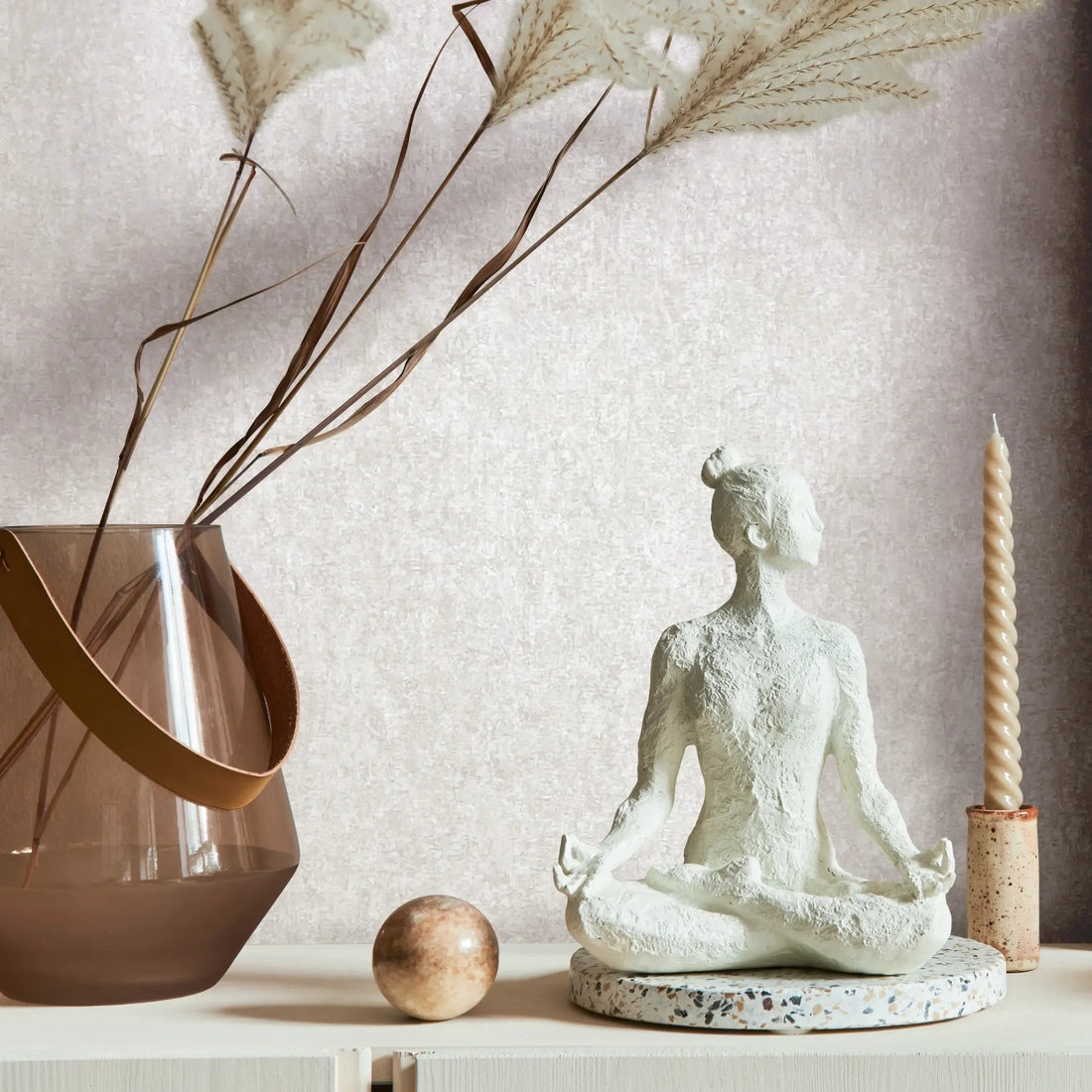 Marburg 35140 wallpaper lounge feature wall behind a console styled with glass vase of dried grasses, seated yoga sculpture, twisted taper candle and stone sphere, creating a calm minimalist mood.