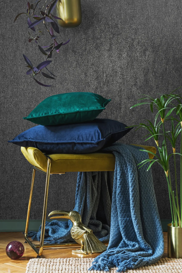 Marburg 35142 wallpaper lounge feature wall with textured dark surface behind a velvet stool topped with blue and green cushions, chunky throw, brass accents and a potted plant, cozy modern vibe.