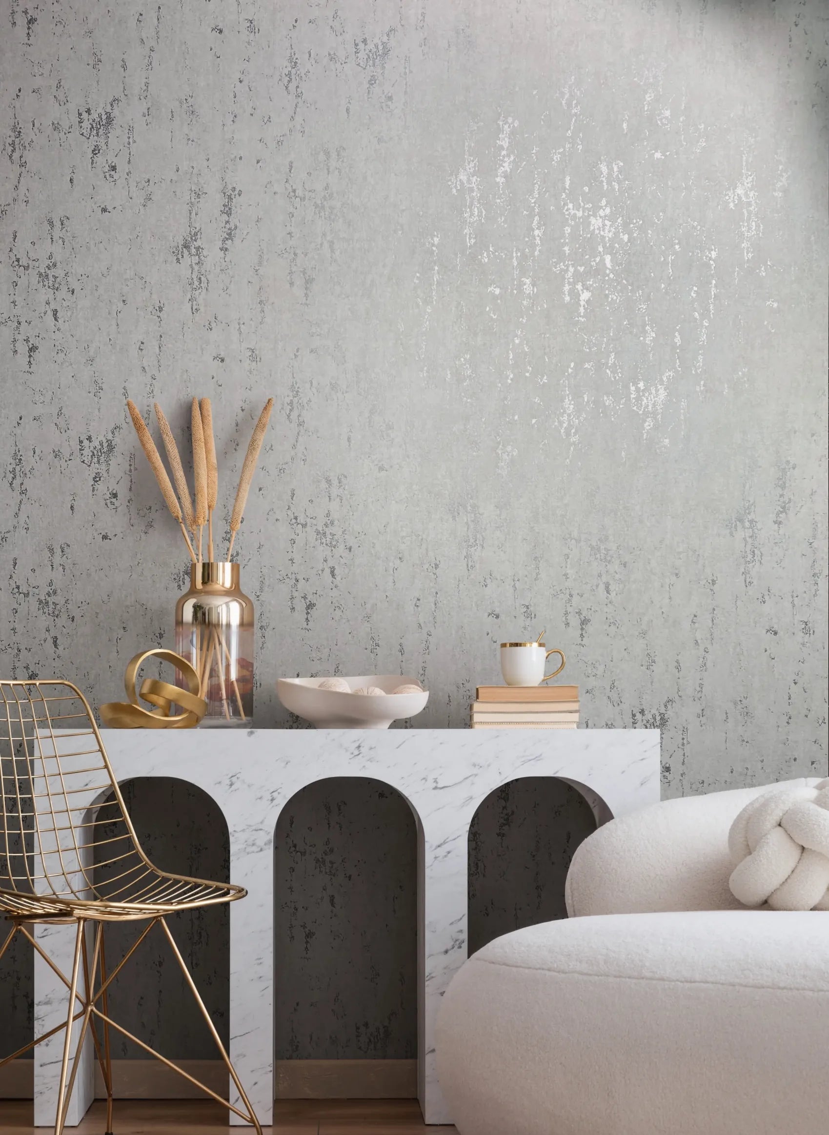 Marburg 35144 wallpaper lounge feature wall behind a marble arch console with dried stems, gold accents, wire chair, and rounded white sofa, calm contemporary mood.