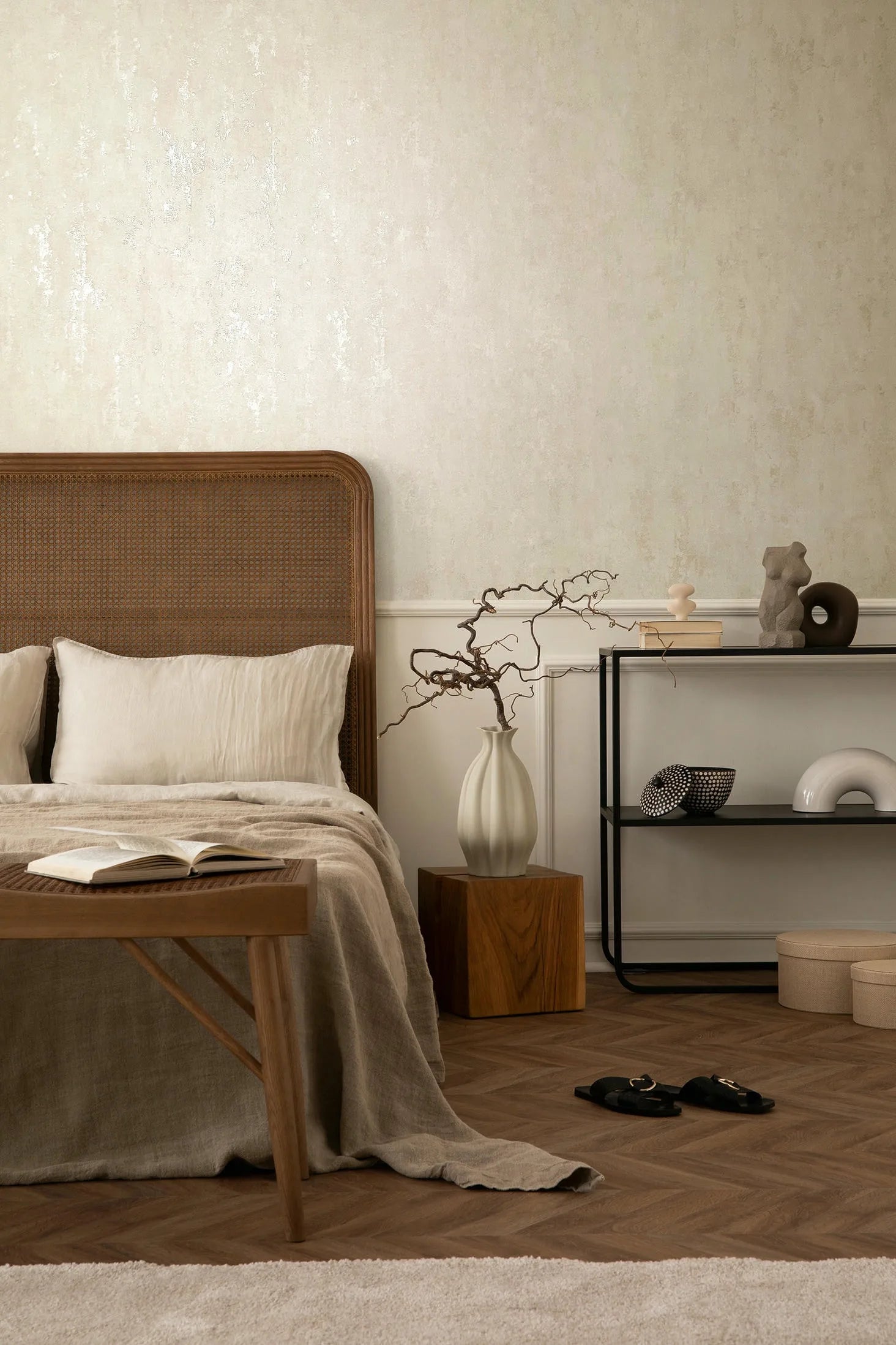 Marburg 35145 wallpaper serene bedroom feature wall behind a cane headboard, linen bedding, wood bench, sculptural vase on a block table, and minimal metal shelving.