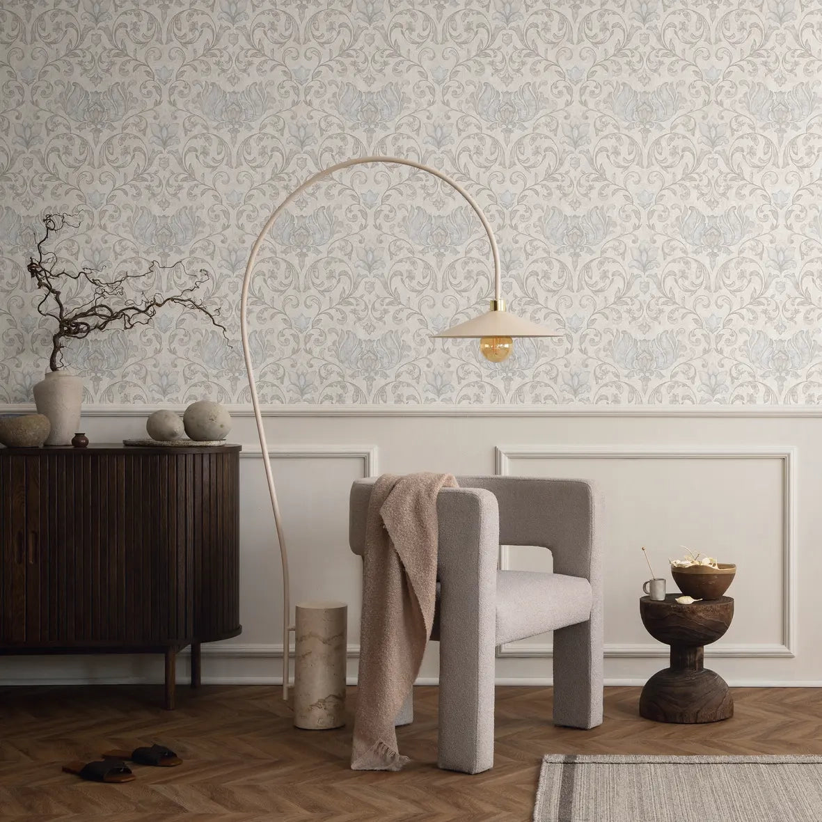 Marburg 35147 wallpaper lounge feature wall above panel moulding, sculptural armchair and arched floor lamp, dark wood sideboard with vases, small wooden side table with bowl and cup, relaxed modern vibe.