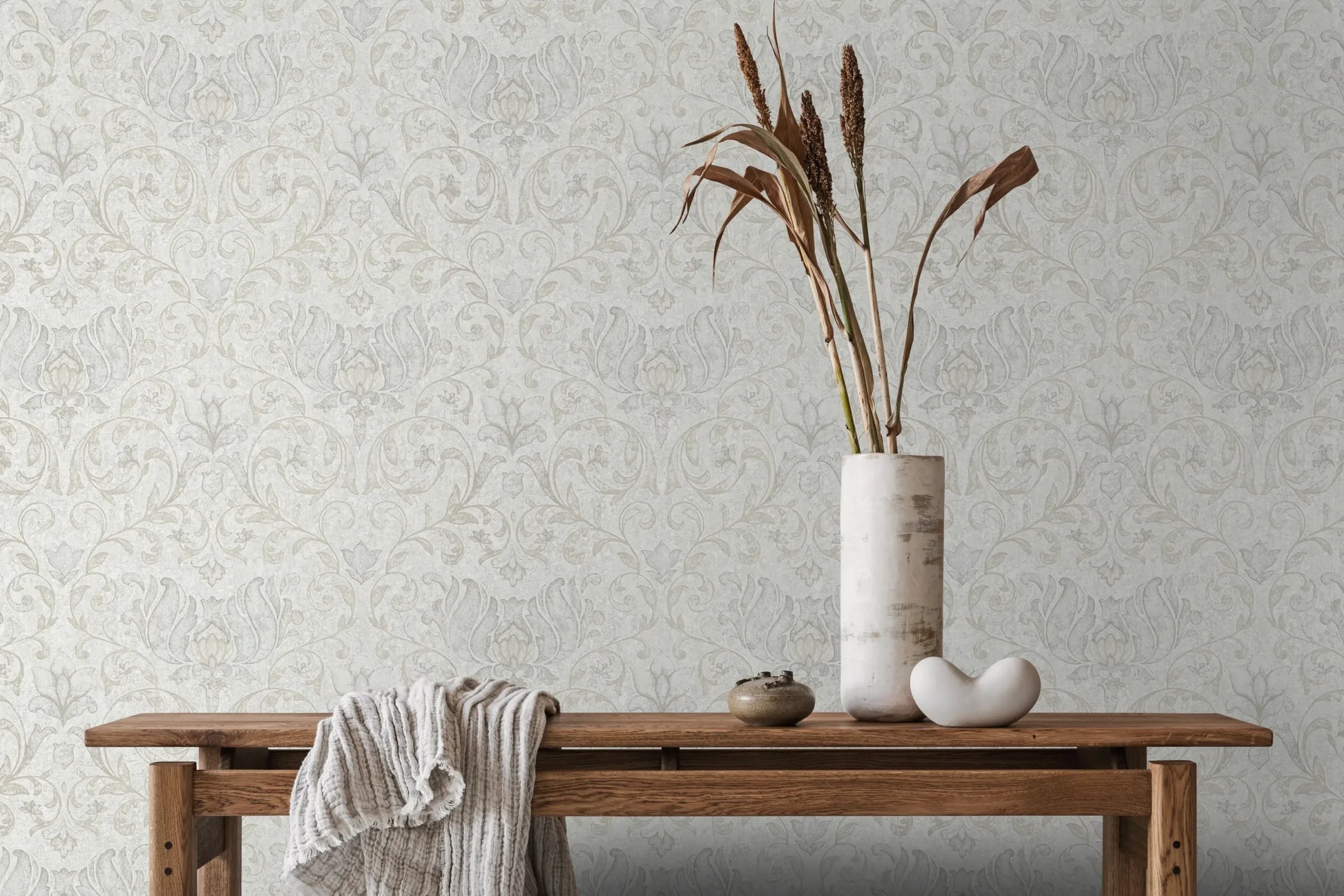Marburg 35148 wallpaper lounge feature wall with subtle damask pattern, wooden console table, draped throw, tall vase of dried stems, small bowl and smooth ornament, serene minimalist mood.