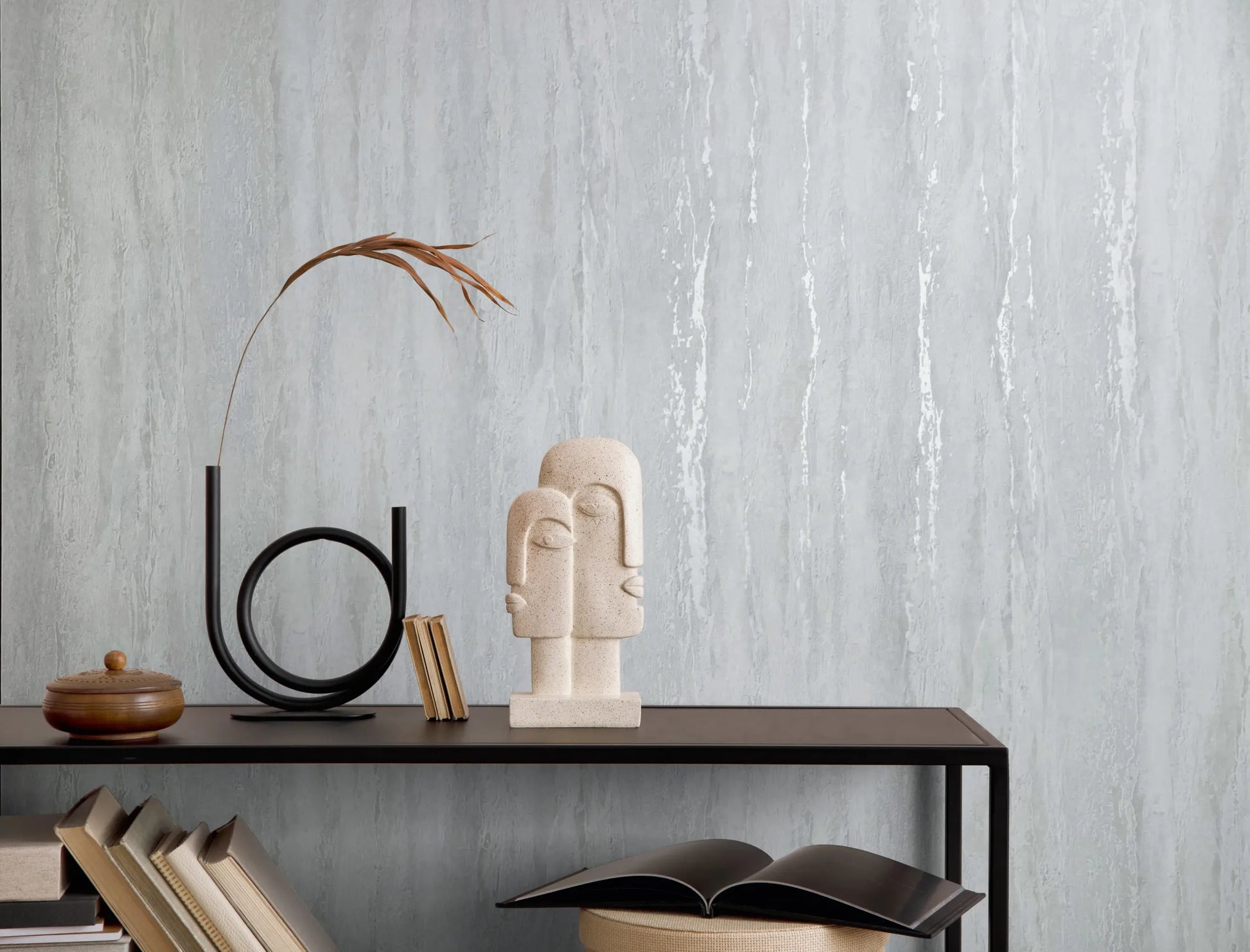 Marburg 35153 wallpaper lounge feature wall with a slim black console table, abstract stone face sculpture, circular metal decor with dried stem, books, and a serene minimalist mood.