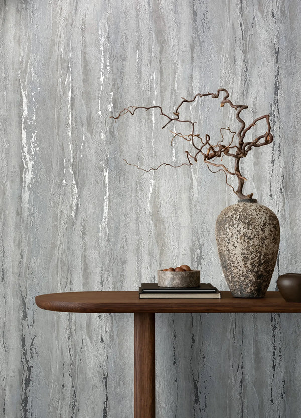 Marburg 35160 wallpaper on a lounge feature wall behind a wooden console table with a textured vase holding sculptural branches, stacked books and a small bowl, creating a calm minimalist mood.