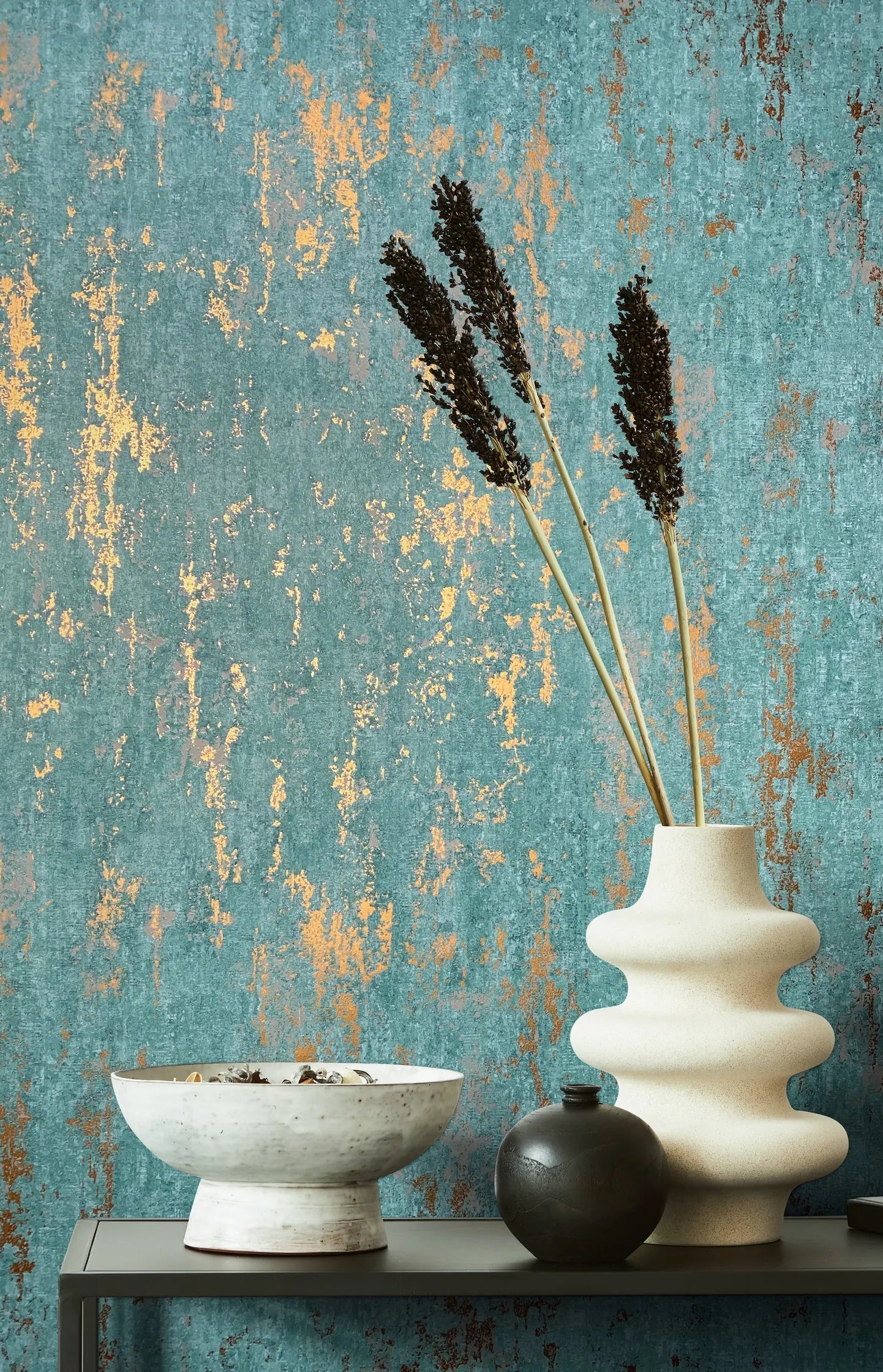 Marburg 35164 wallpaper lounge feature wall with distressed teal and warm metallic texture behind a slim console holding a sculptural cream vase with dried stems, a dark round vase, and a wide ceramic bowl for a calm.