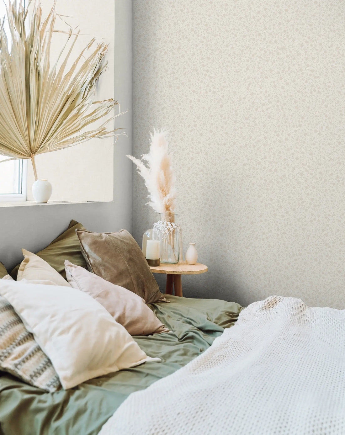 Marburg 47601 wallpaper in a serene bedroom feature wall behind a low bed piled with pillows and a knit throw, round bedside table with pampas in a glass vase, candle and small vase beside a window.