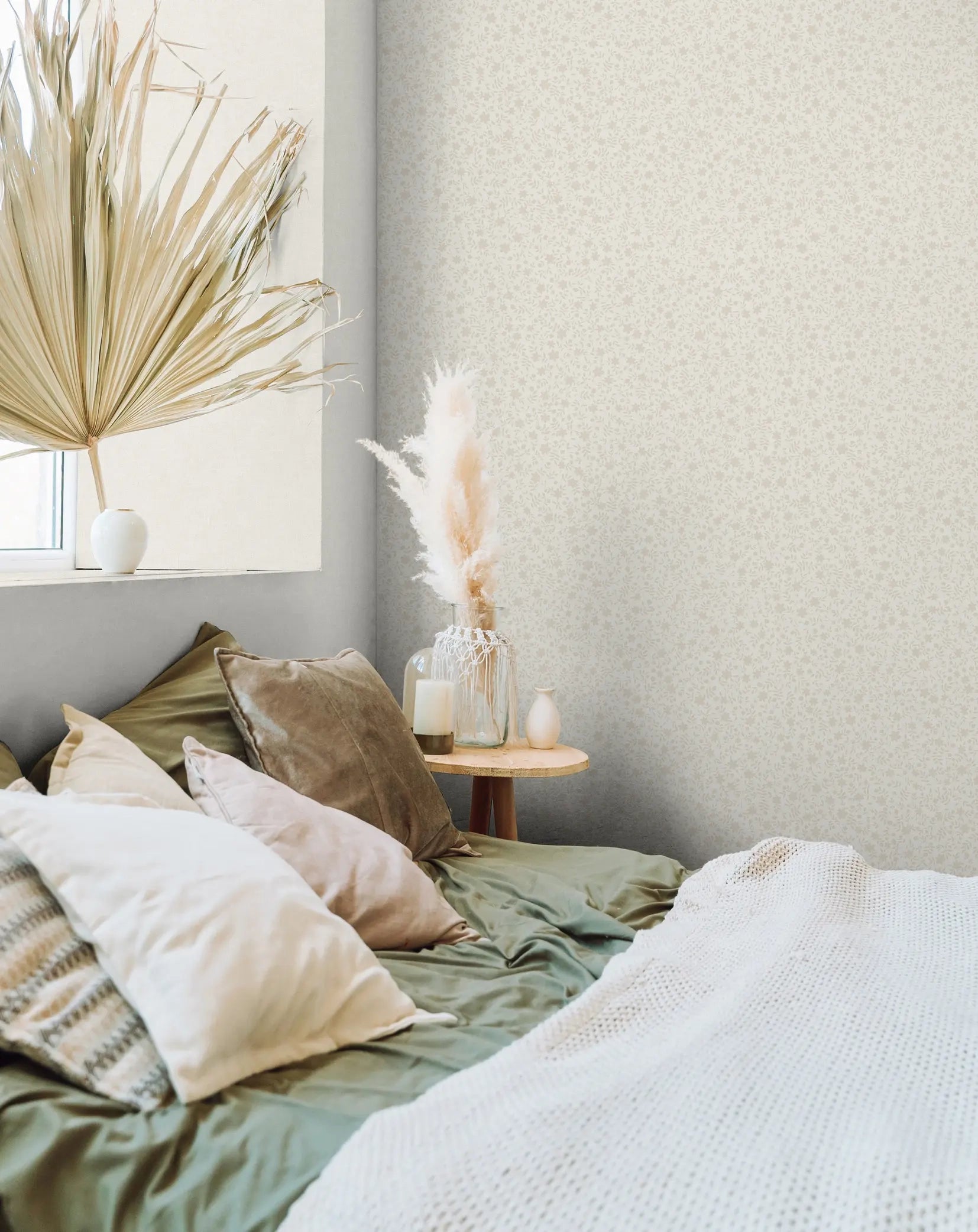 Marburg 47601 wallpaper in a serene bedroom feature wall behind a low bed piled with pillows and a knit throw, round bedside table with pampas in a glass vase, candle and small vase beside a window.