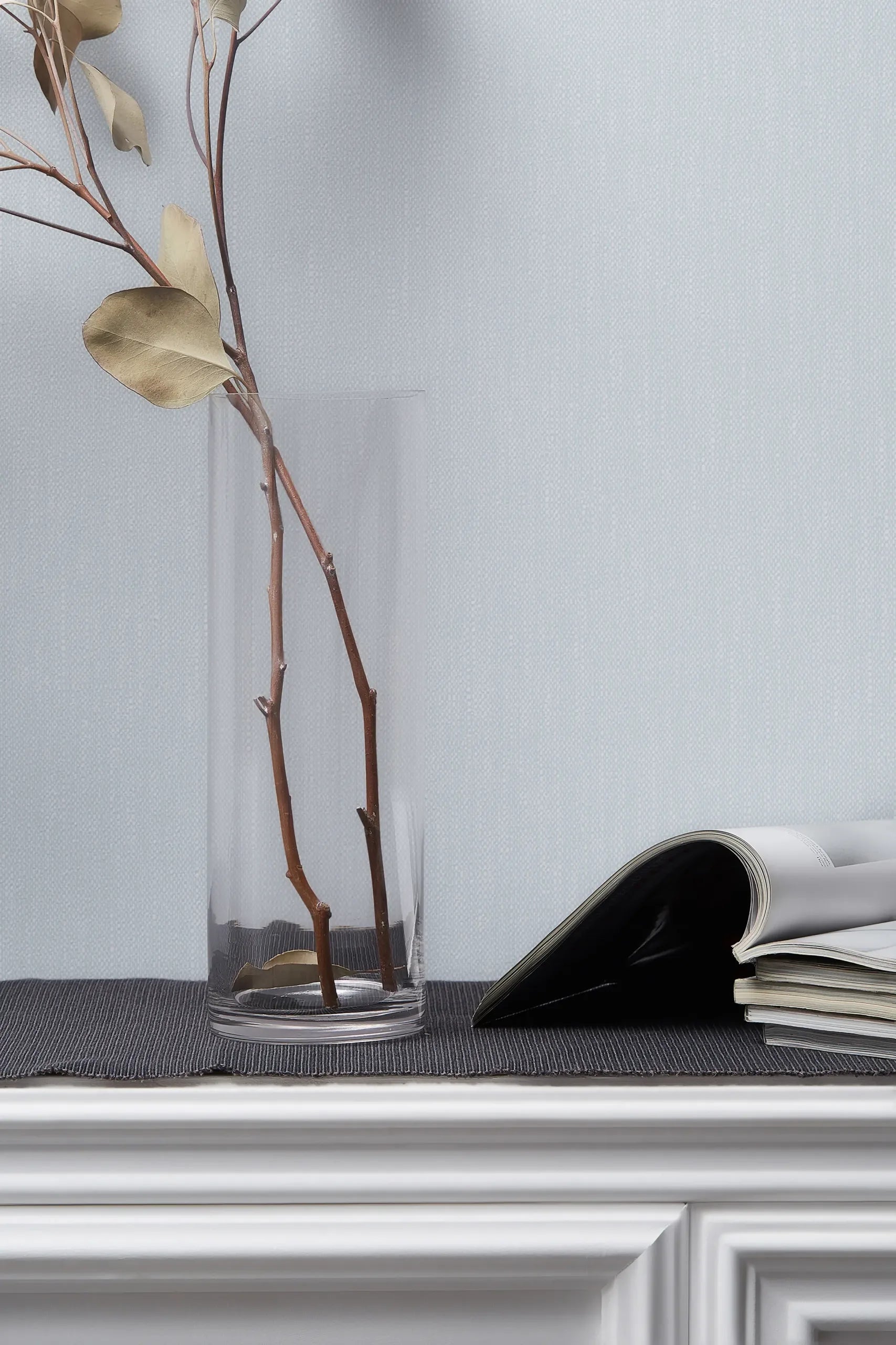 Marburg 47609 wallpaper lounge feature wall behind a white paneled console with a glass vase of dried branches and open magazines, calm minimalist mood.