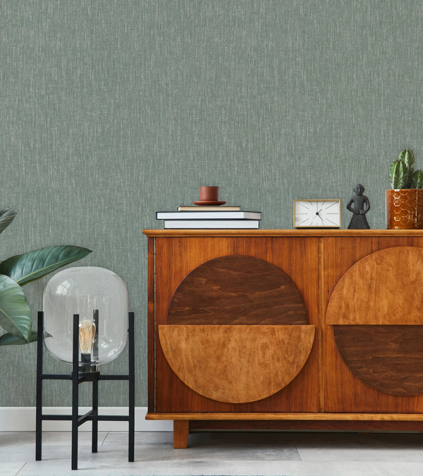 Marburg 47615 wallpaper lounge feature wall behind a mid-century wood sideboard with books and candle, small clock and figurine, cactus planter, glass floor lamp and leafy plant, calm modern feel.