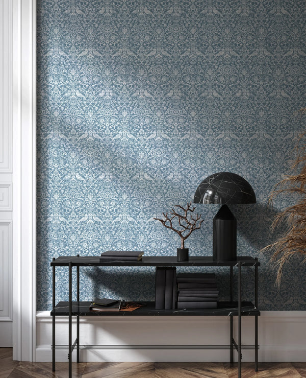 Marburg 47623 wallpaper lounge feature wall behind a slim black console with stacked books, a sculptural branch and a dome lamp, herringbone wood floor, serene modern mood.