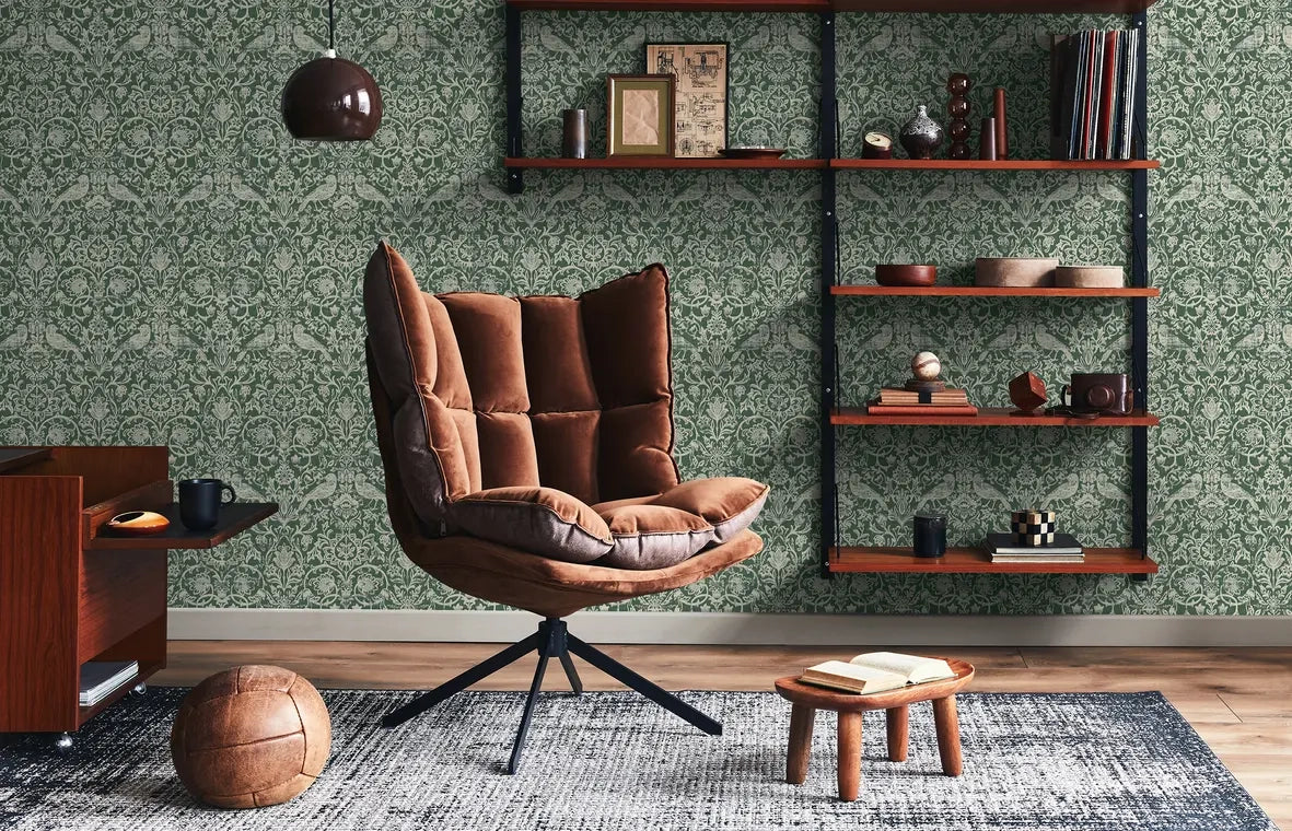 Marburg 47624 wallpaper lounge feature wall behind brown cushioned swivel chair, floating wood shelves with decor, small stool with open book, pendant light and textured rug for a cozy mid-century feel.