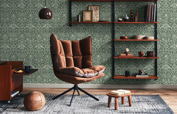 Marburg 47624 wallpaper lounge feature wall behind brown cushioned swivel chair, floating wood shelves with decor, small stool with open book, pendant light and textured rug for a cozy mid-century feel.