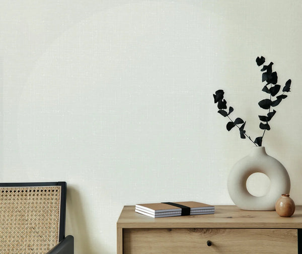 Marburg 47630 wallpaper lounge feature wall with subtle texture, rattan chair and wood sideboard topped with a sculptural round vase, dark stems, small bud vase and notebooks, calm minimalist mood.
