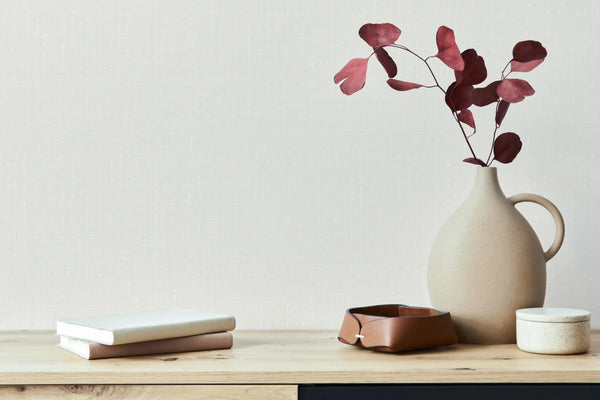 Marburg 47631 wallpaper lounge feature wall with a wooden console, ceramic jug vase holding burgundy leaves, stacked books, leather catchall, and a small lidded pot for a calm minimalist feel.