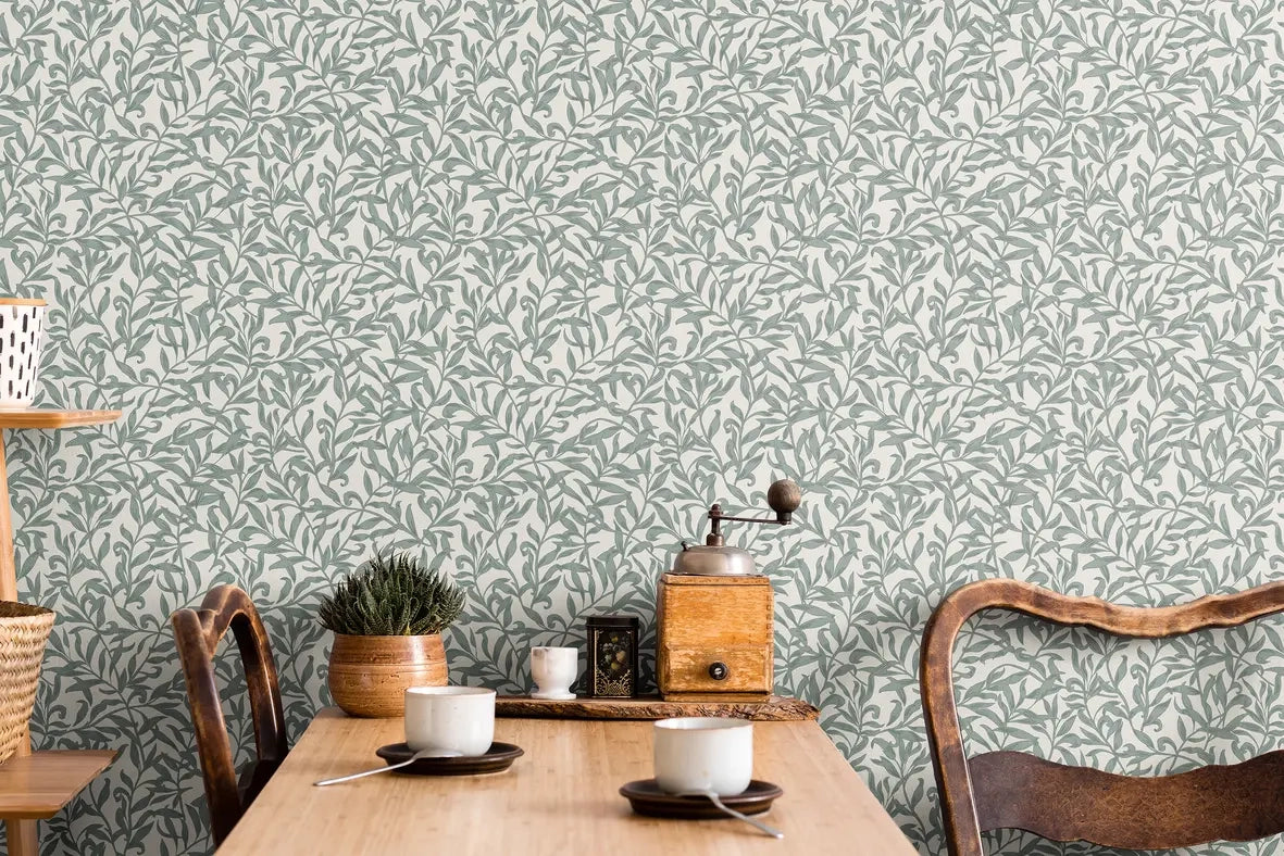 Marburg 47639 wallpaper lounge feature wall with leafy botanical pattern behind a rustic wooden table and worn chairs, cups and saucers, a small potted plant and vintage coffee grinder, relaxed cozy mood.