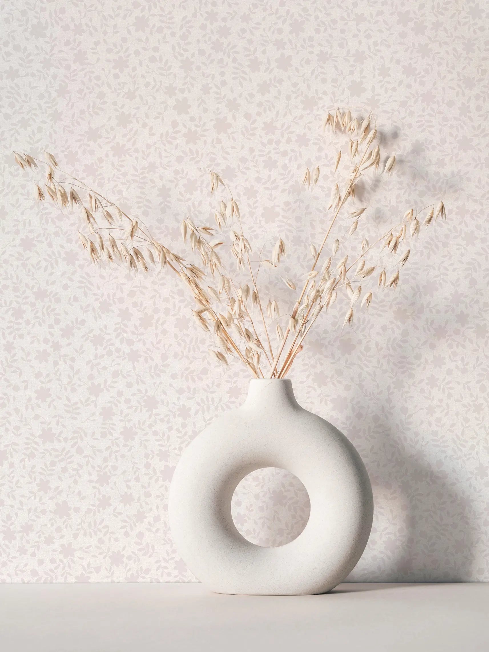 Marburg 47644 wallpaper lounge feature wall with delicate floral pattern behind a sculptural ceramic ring vase holding dried stems, calm minimalist mood.