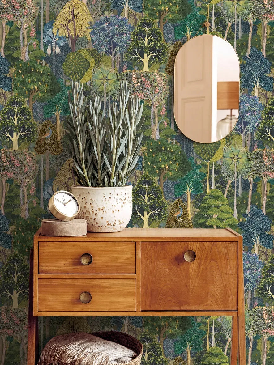Marburg 47650 wallpaper lounge feature wall with lush forest print behind a wooden console table, oval mirror, potted plant and clock, basket with throw, cozy natural feel.