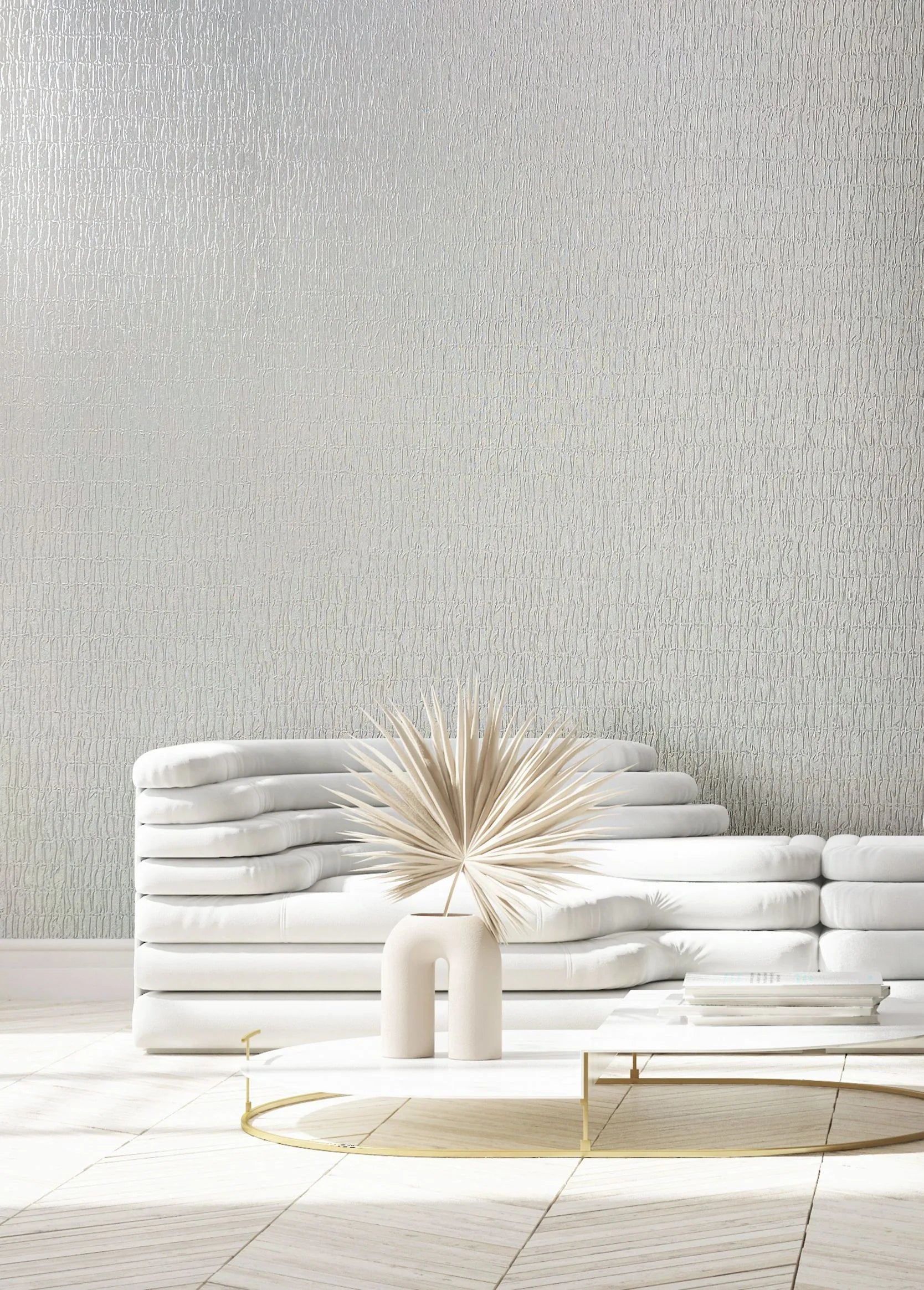 Marburg Kumano 34501 White wallpaper with a subtle vertical organic texture shown behind a low white sofa and a round coffee table holding a sculptural vase with dried palm leaves and a small stack of books.
