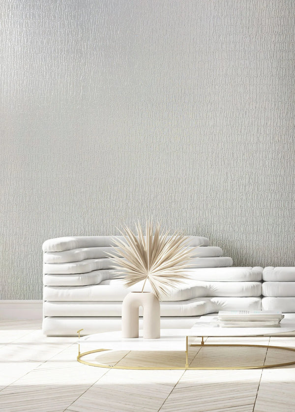 Marburg Kumano 34501 White wallpaper with a subtle vertical organic texture shown behind a low white sofa and a round coffee table holding a sculptural vase with dried palm leaves and a small stack of books.