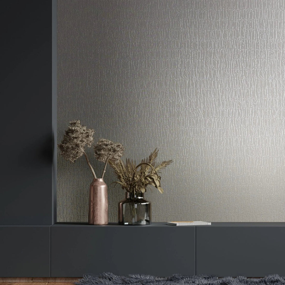 Marburg Kumano 34503 Gray wallpaper with a subtle irregular linear texture shown as a feature wall behind a dark cabinet, with two vases holding dried stems, a book, a shag rug, and a wood floor in view.