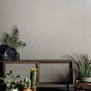 Marburg Kumano 34504 Light gray textured wallpaper behind a cane-front console with black dishes, a glass bottle with branches, potted cacti and succulents, a round woven rug, and part of a wooden chair.