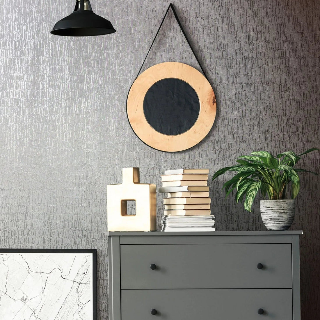 Marburg Kumano 34506 Room vignette showing gray textured wallpaper with an irregular vertical pattern behind a gray three-drawer dresser. A round hanging mirror and a black pendant light are on the wall, with a potted plant, stacked books, and a geometric sculpture on the dresser.