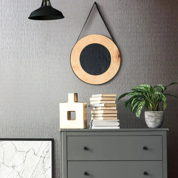 Marburg Kumano 34506 Room vignette showing gray textured wallpaper with an irregular vertical pattern behind a gray three-drawer dresser. A round hanging mirror and a black pendant light are on the wall, with a potted plant, stacked books, and a geometric sculpture on the dresser.